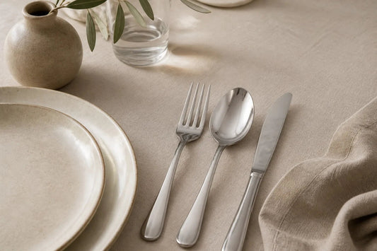 Polished 18/10 stainless steel fork, spoon and knife laid on a natural linen tablecloth beside a cream ceramic plate